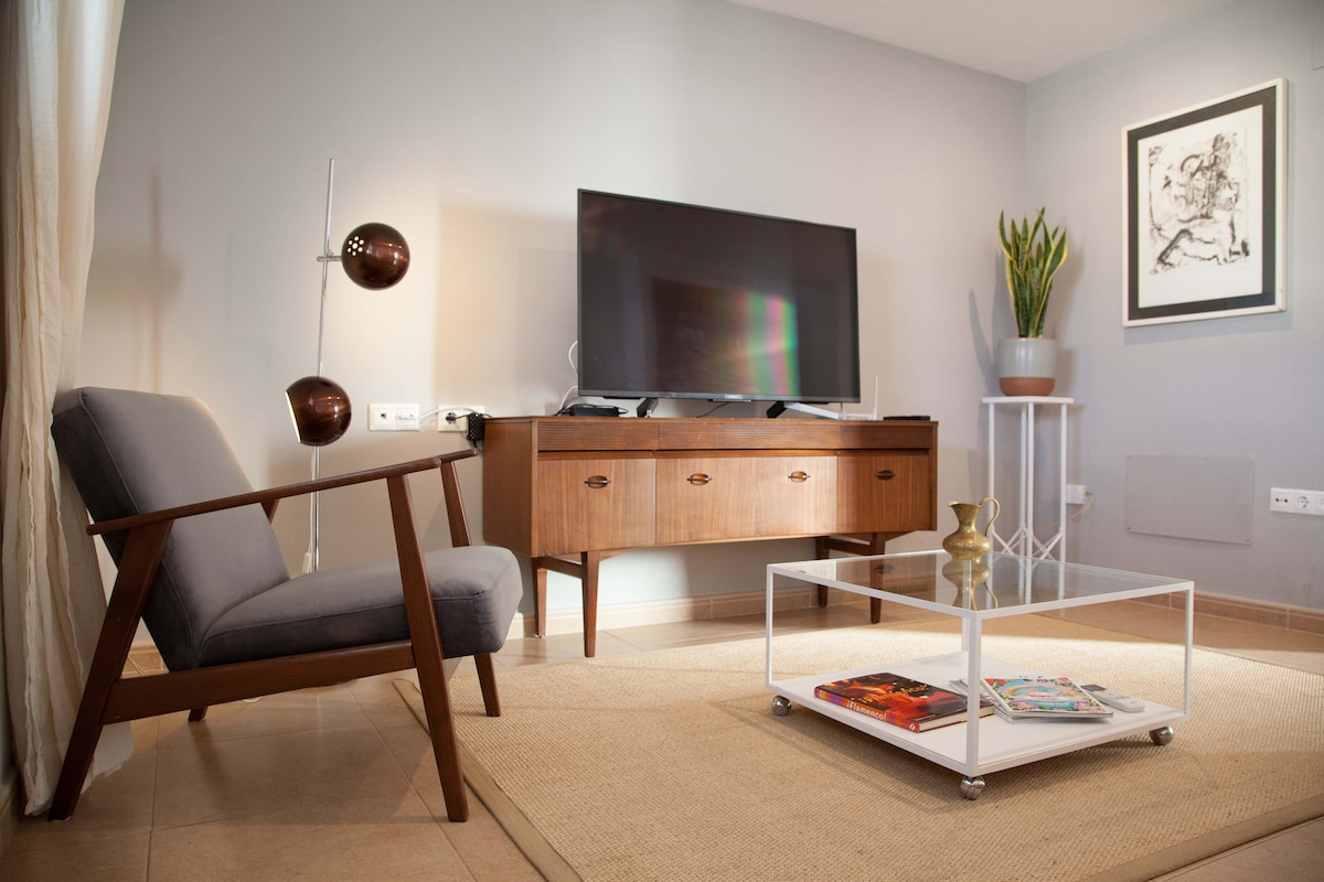 A modern living area features a wooden coffee table displaying magazines, complemented by a stylish gray armchair. A flat-screen television is mounted on a mid-century style media console. Natural light filters through the window beside a decorative plant placed on a small side table.