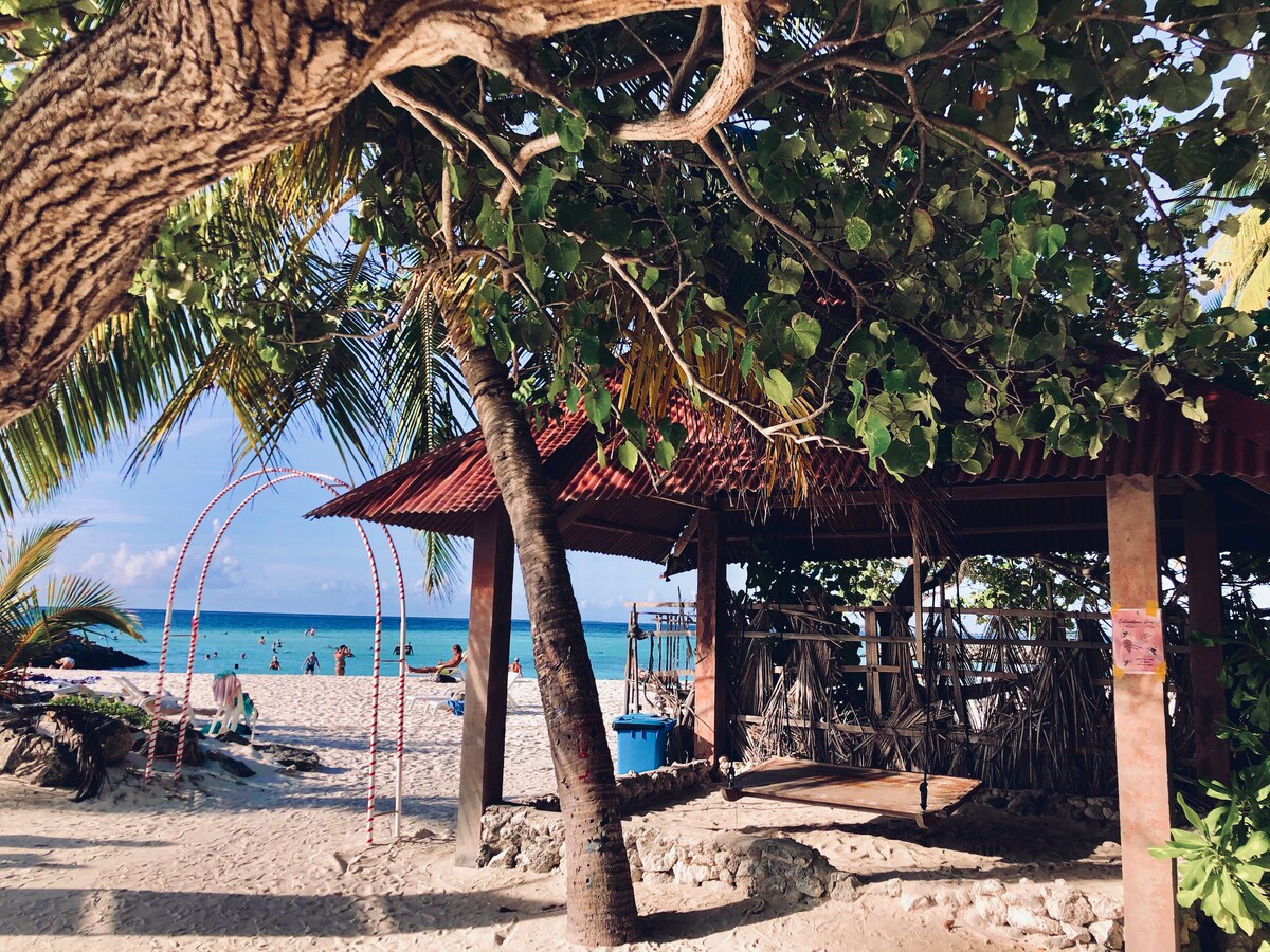 A shaded pavilion is surrounded by lush green trees and a sandy beach. The sun shines on the blue waters in the background, while beachgoers enjoy the space. Decorative arches can be seen nearby, adding character to the coastal view.