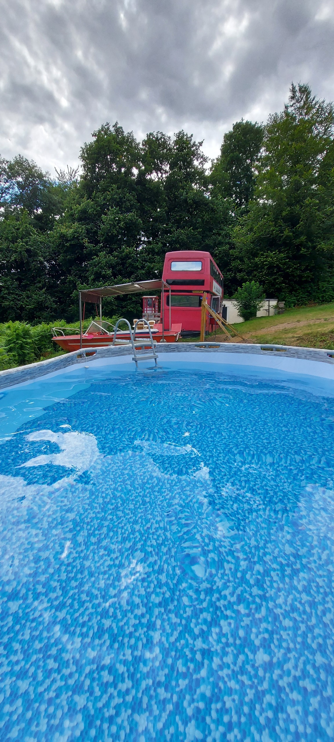 A circular swimming pool is shown, featuring clear blue water and a ladder inviting access. In the background, a red structure is positioned near the pool, surrounded by greenery and tall trees, contributing to a tranquil outdoor setting.