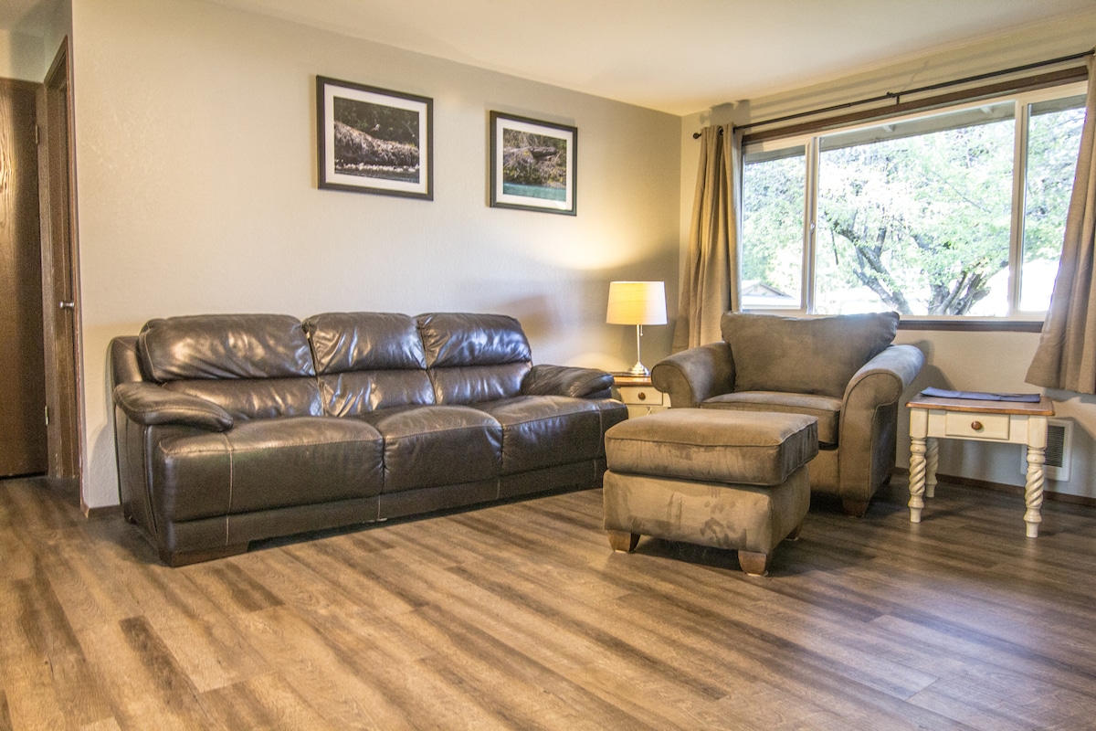 A welcoming living area features a dark leather sofa paired with a comfortable armchair. A side table holds a lamp, and natural light filters through the large window adorned with curtains, illuminating the room and highlighting the wood-like flooring.