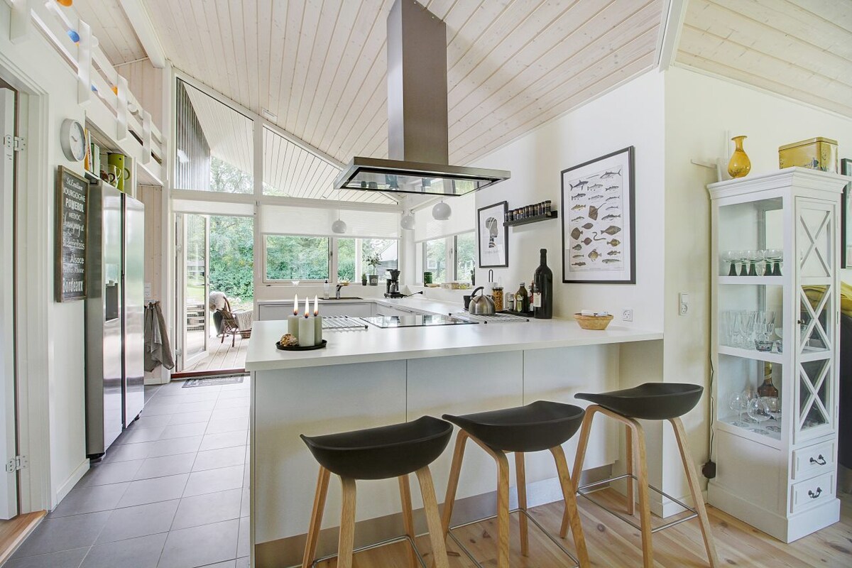 A bright kitchen area features a spacious island with seating for three, complemented by modern appliances and a sleek hood above the cooktop. Large windows provide natural light, while the open layout connects to a view of the outdoor terrace.