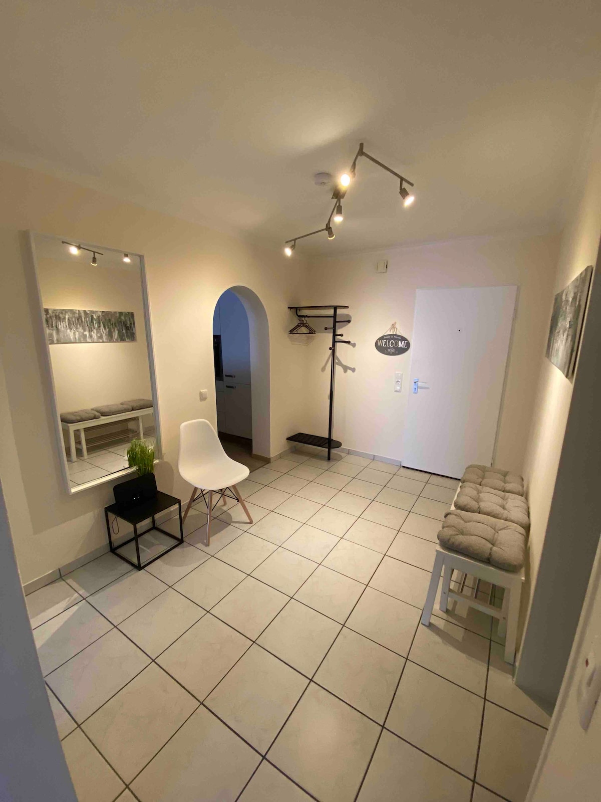A bright entryway features tiled flooring and is illuminated by recessed lighting. A mirror and a decorative piece are mounted on the wall. A white chair and a small black table sit beside a cushioned bench, while a coat rack is positioned to the side.