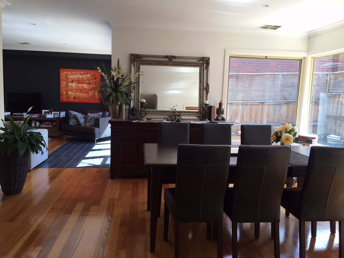An elegant dining area features a large, dark wood table surrounded by eight upholstered chairs. Natural light floods in through expansive windows, highlighting wooden flooring. A decorative mirror reflects the ambiance, while a potted plant and floral arrangements add subtle touches.
