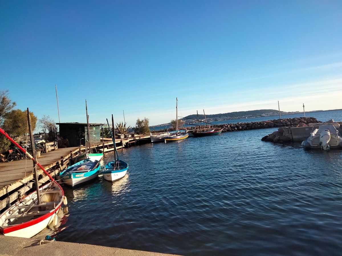 A serene waterfront scene shows several fishing boats moored alongside a wooden dock. Calm blue waters reflect the clear blue sky, while distant hills are seen along the horizon. The area appears peaceful, with minimal activity on the water.