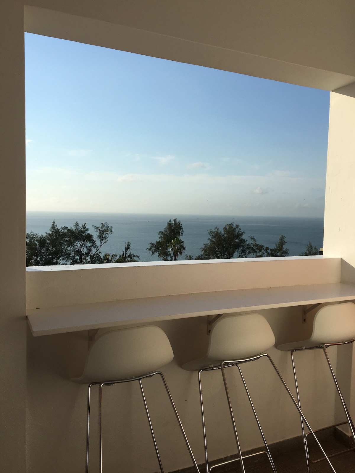 A bright window frame offers an unobstructed view of the ocean horizon, complemented by a white countertop. Modern white bar stools are positioned along the counter, creating an inviting space to enjoy the expansive scenery.