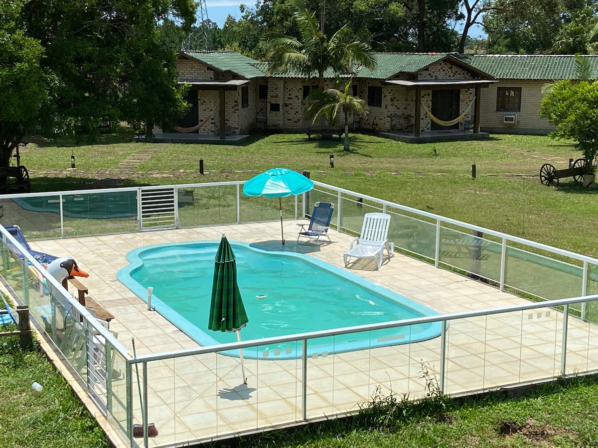 A sunlit pool area is enclosed by a safety fence, featuring a refreshing turquoise pool. Sun loungers and an umbrella provide shade, while green trees frame the picturesque surroundings. In the background, spacious cabins are visible, offering a serene retreat.