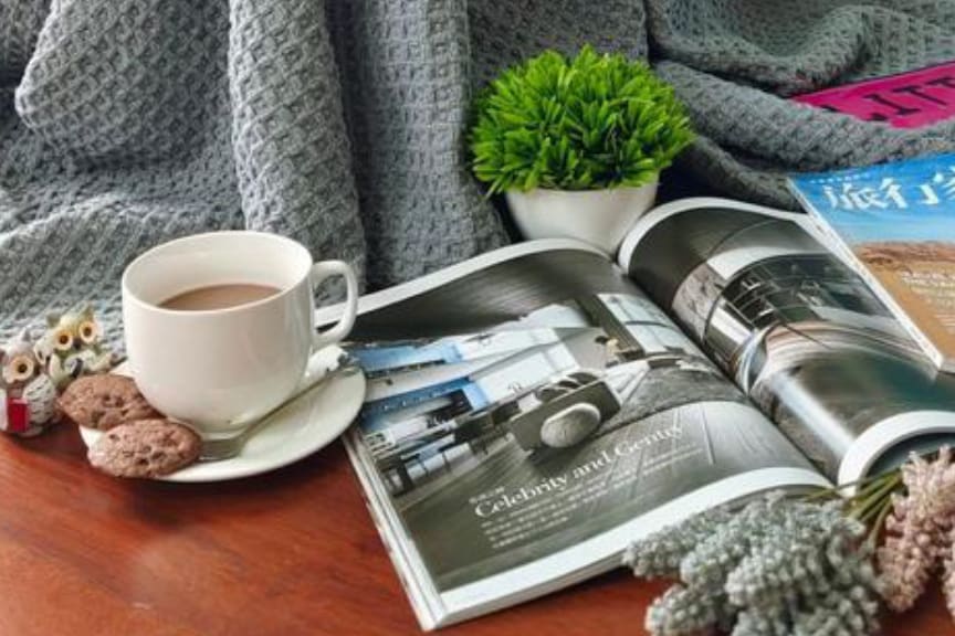 A coffee cup sits next to a magazine on a wooden table, accompanied by a small plant and cookies. A cozy blanket partially covers the table, offering a comfortable setting for relaxation and leisure.