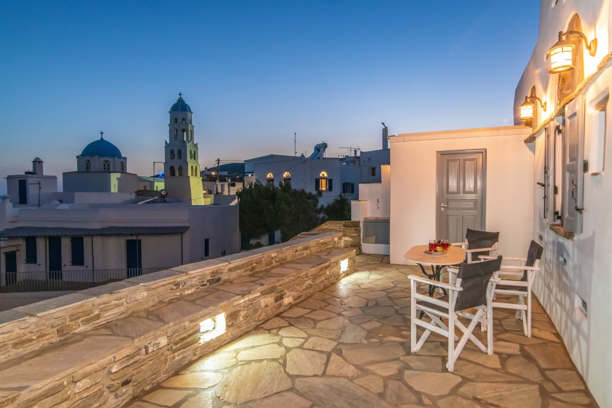 A stone terrace is presented with two chairs and a small table, illuminated softly by outdoor lighting. The background features traditional Cycladic architecture, including a blue-domed church, under a twilight sky transitioning to evening shades.