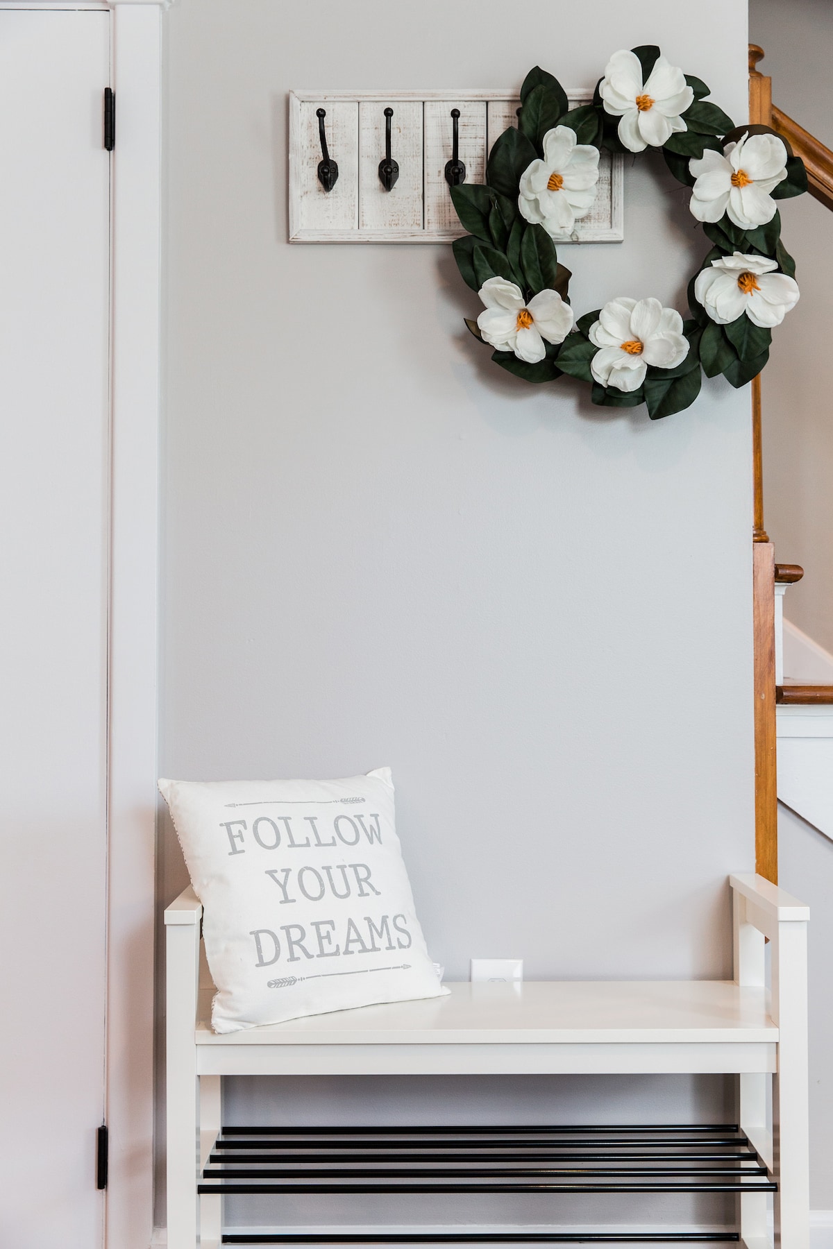 A welcoming entryway features a light-colored bench with a striped shelf underneath. A decorative wreath with white flowers is mounted on the wall above four hooks. A cushion with 'FOLLOW YOUR DREAMS' text rests on the bench, adding a subtle personal touch.