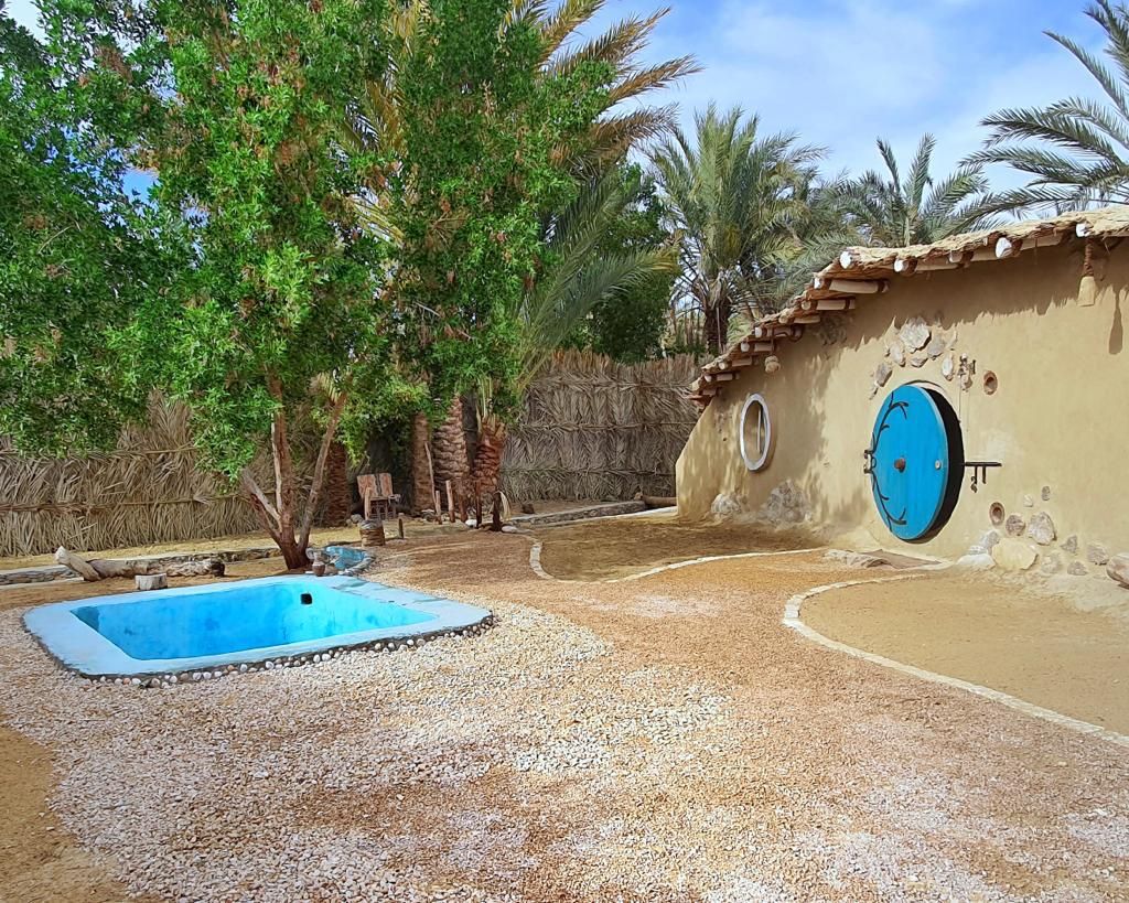 A serene outdoor space is featured, with a small turquoise pool framed by a pebbled pathway. The sunlit area is surrounded by lush palm trees and rustic walls made of natural materials, complemented by a unique round door colored in blue.