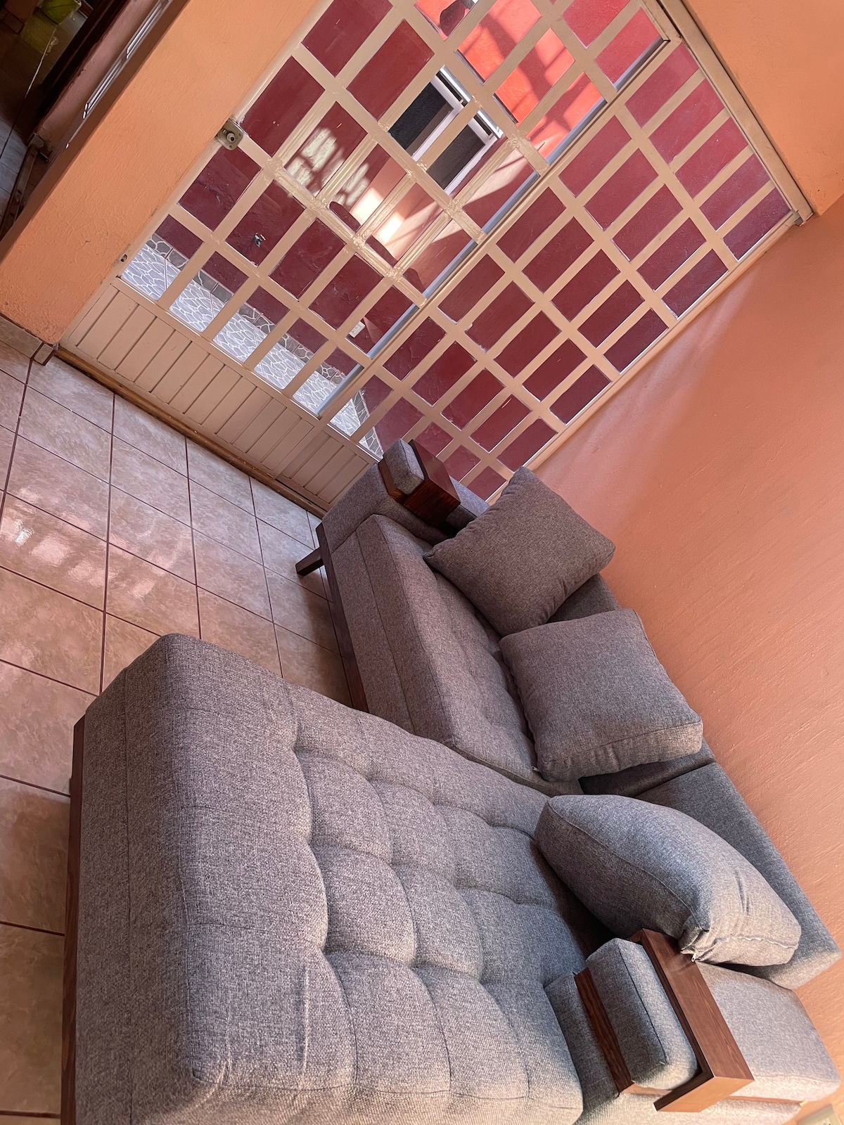 A gray sectional sofa is positioned in the room, featuring plush cushions and wooden armrests. Natural light filters through a door with a grid design, partially illuminating the glossy tile floor in a warm tone.