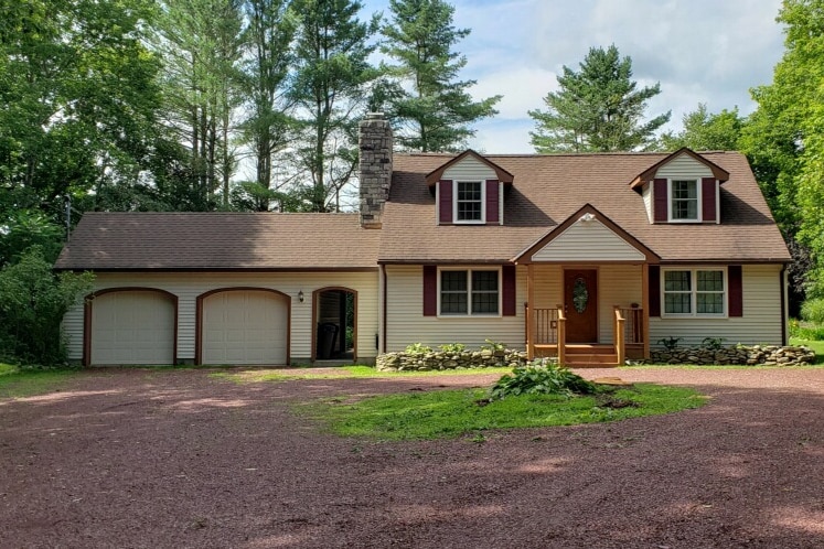 A charming exterior view of the house is displayed, featuring a welcoming entrance with a front porch and wooden steps. The property is flanked by a two-car garage, with a gravel driveway leading up to the house. Lush greenery surrounds the building, providing a sense of seclusion.