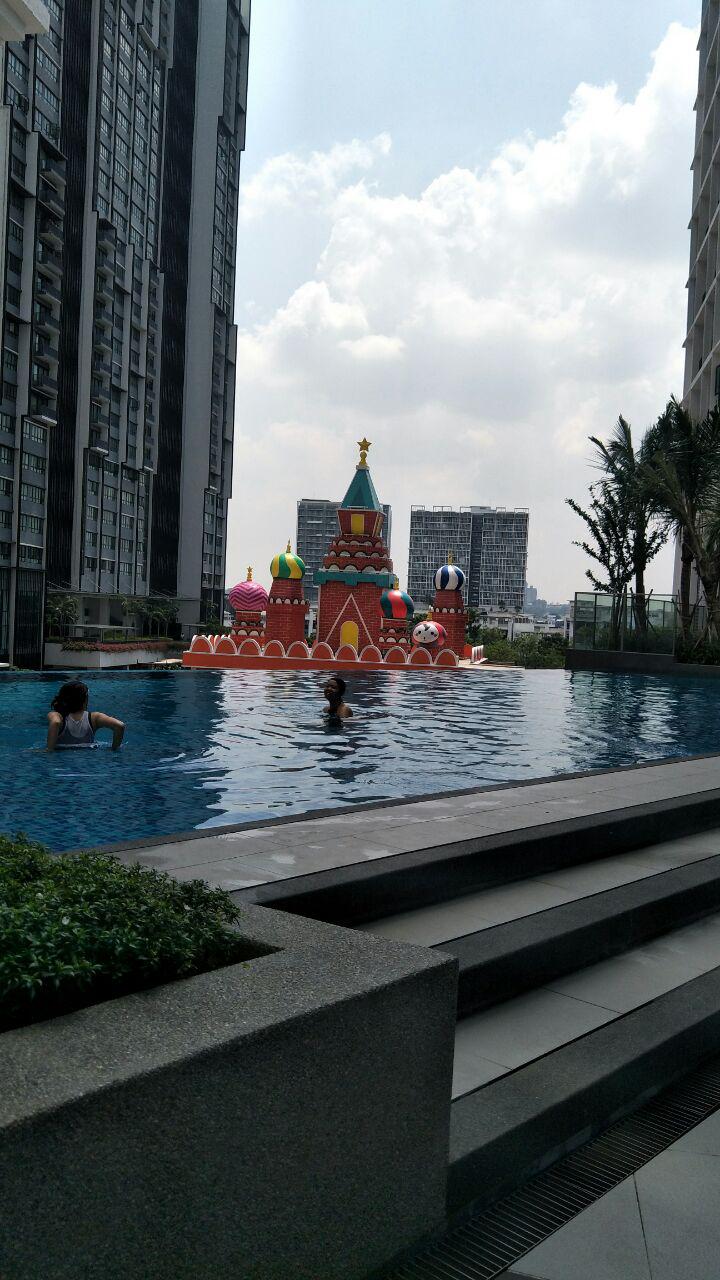 A swimming pool area is highlighted with a colorful floating structure resembling a castle, surrounded by lush greenery. The pool is bordered by modern apartment buildings, with a clear blue sky and scattered clouds in the background, creating a serene atmosphere for relaxation.