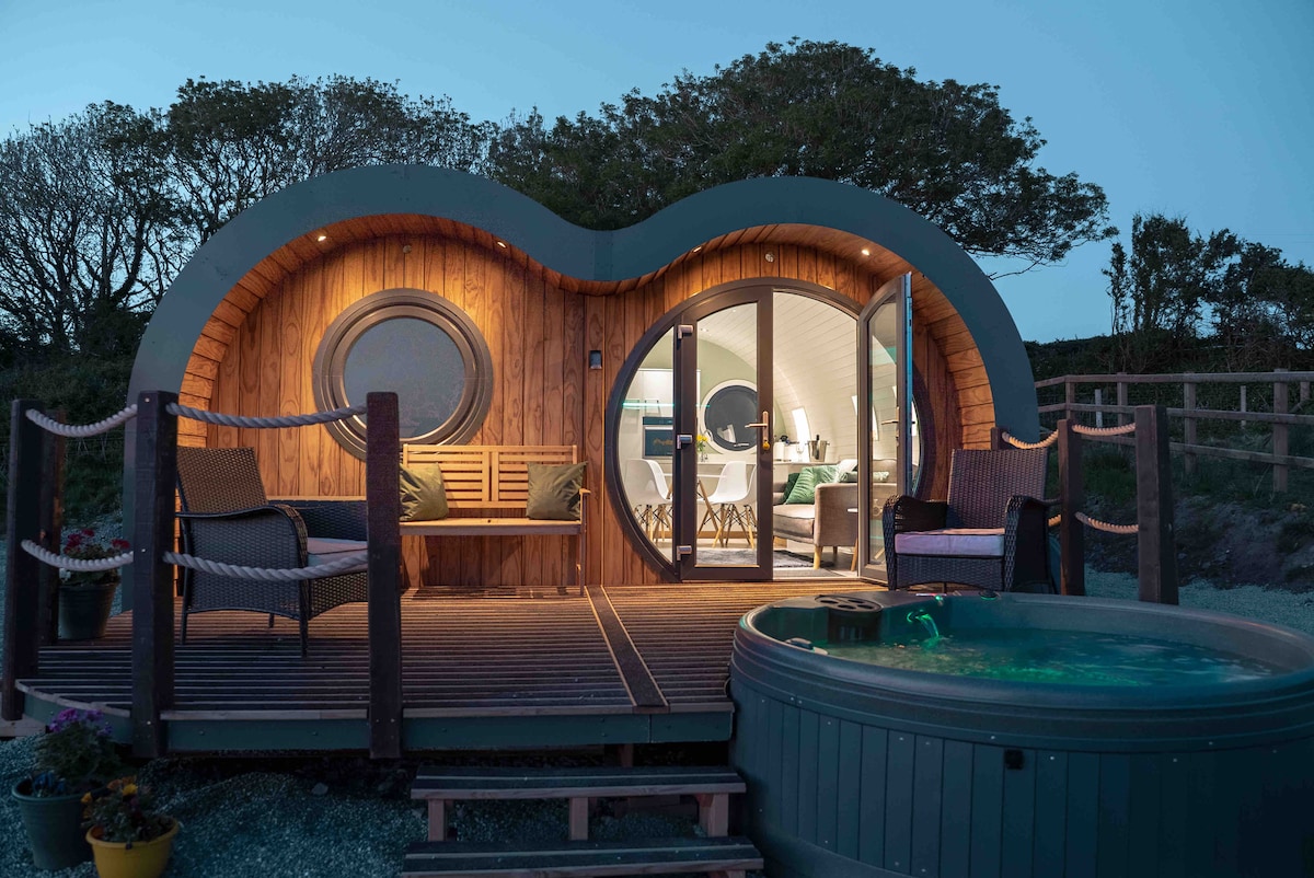 The unique exterior of a luxury glamping accommodation is highlighted, featuring round windows and wooden accents. A spacious deck surrounds the structure, with two comfortable chairs positioned for relaxation. A hot tub sits nearby, creating an inviting atmosphere for enjoying the outdoors.