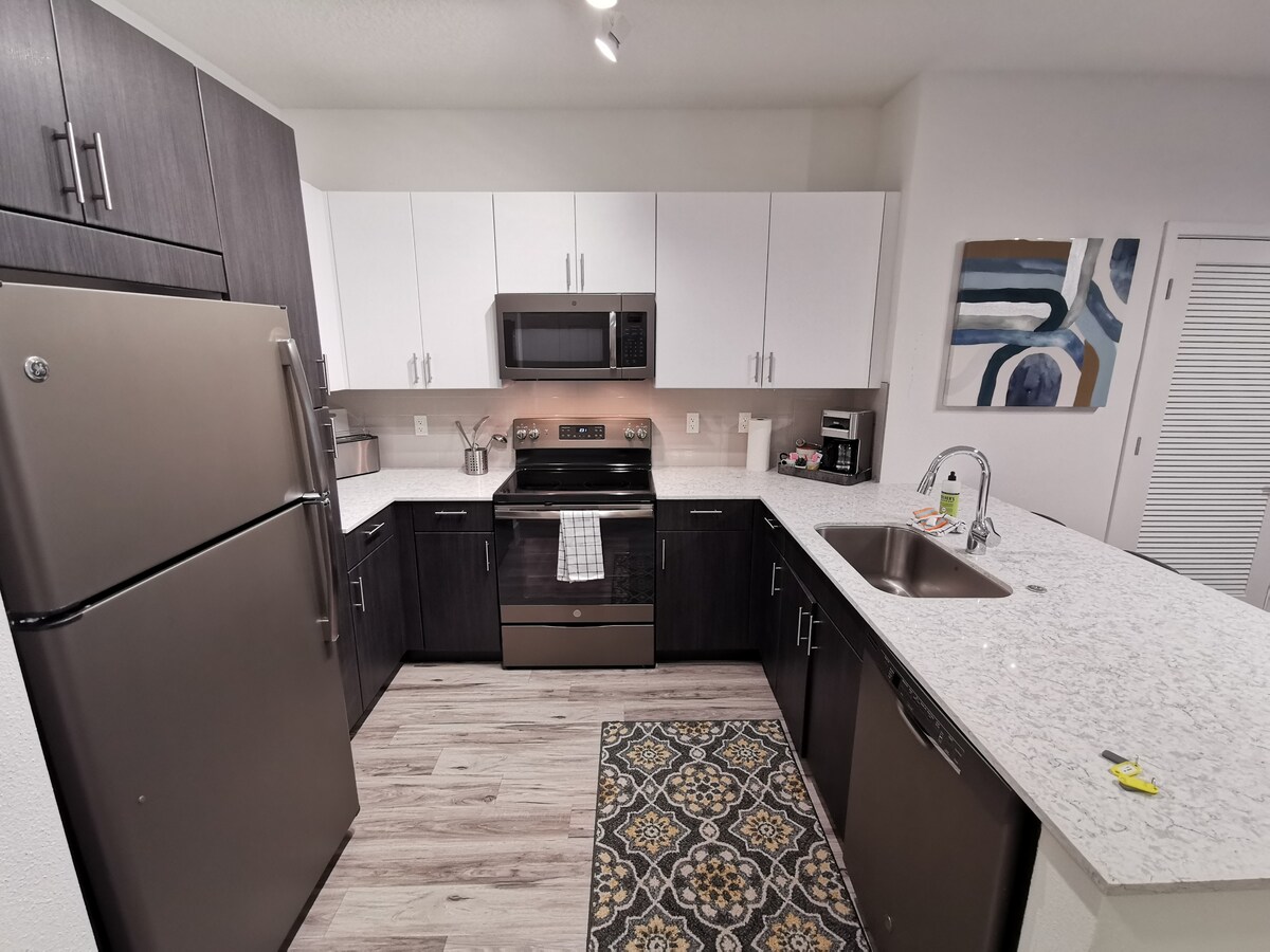 A modern kitchen features dark cabinetry paired with white upper cabinets, complemented by a light granite countertop. Stainless steel appliances, including an oven and refrigerator, are present. A patterned rug adds warmth to the space, which is illuminated by overhead lighting.