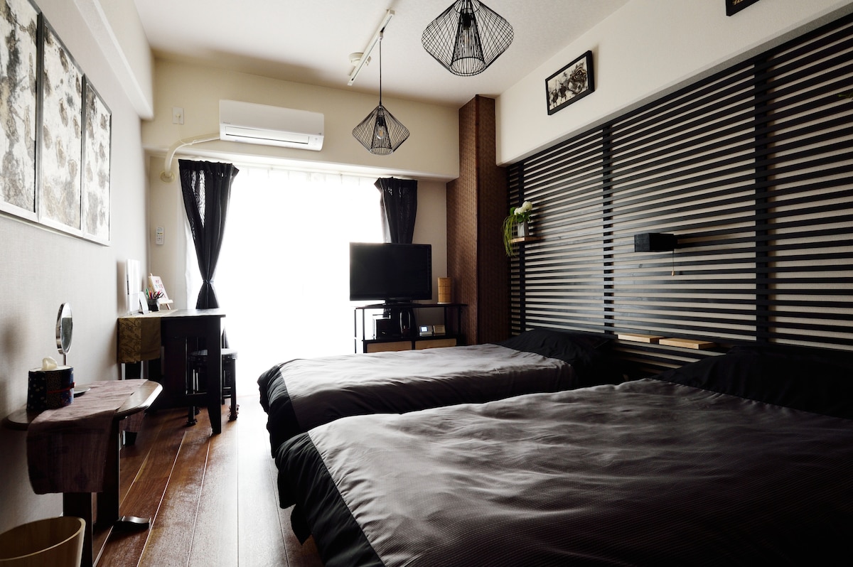 A spacious room features two single beds with dark bedding, positioned side by side. A large window is covered by dark curtains, allowing natural light to filter in. A television is mounted on a wall, and stylish light fixtures hang from the ceiling.