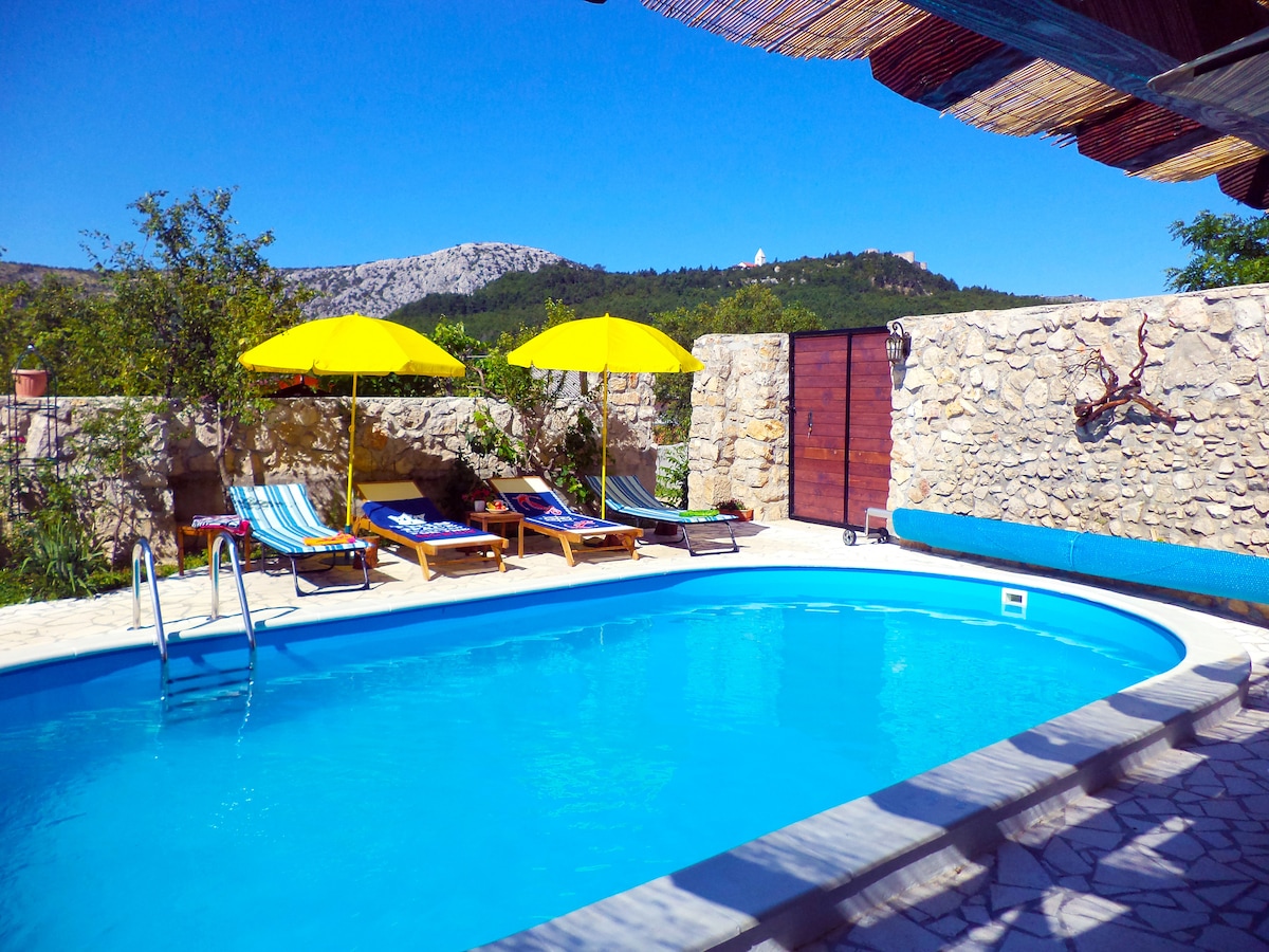 A refreshing pool is positioned within a stone courtyard, surrounded by lush greenery. Two vibrant yellow umbrellas provide shade for the loungers, which include striped cushions. A ladder leads into the circular pool, inviting relaxation under clear blue skies and a backdrop of rolling hills.