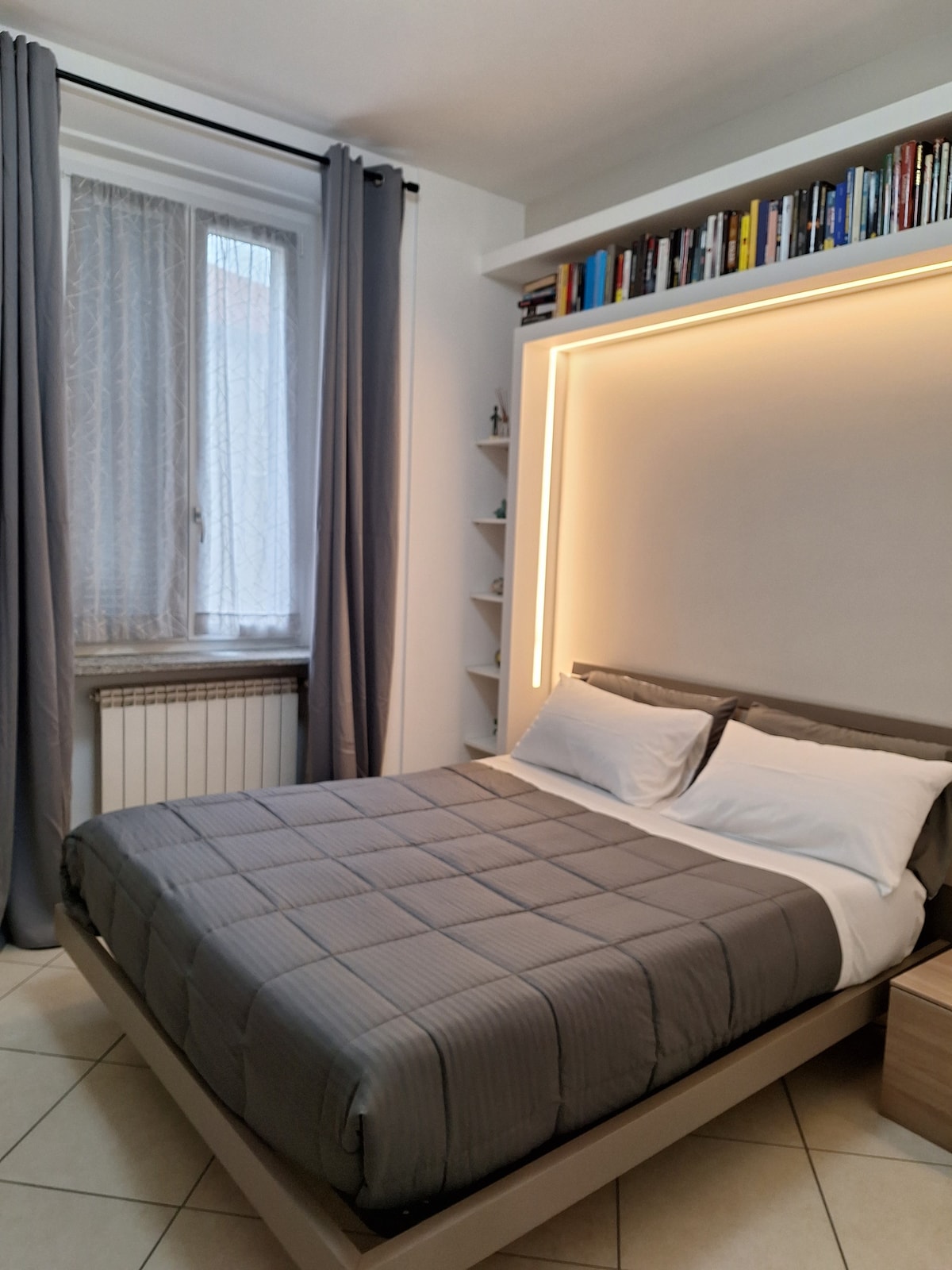 A comfortable sleeping area is presented with a spacious bed dressed in a soft gray coverlet. Light from a nearby window filters through sheer curtains, while a shelf of books is positioned above the bed, adding to the room's inviting design.