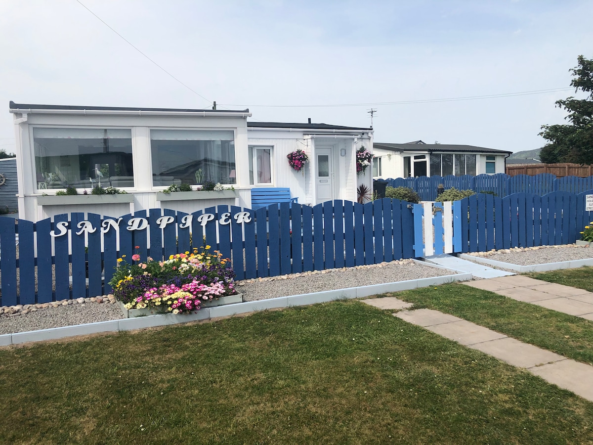 A charming bungalow is set behind a distinctive blue fence adorned with the name 'SANDPIPER.' Colorful flower beds line the front, featuring various blooming plants. The property is complemented by a manicured lawn and a stone pathway leading to the entrance.