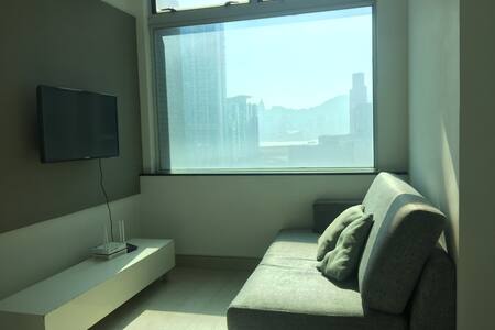 A modern sitting area is highlighted by large windows that provide ample natural light and views of the surrounding skyline. A soft gray sofa is positioned against the wall, accompanied by a small coffee table. A flat-screen television is mounted on the wall opposite the sofa.