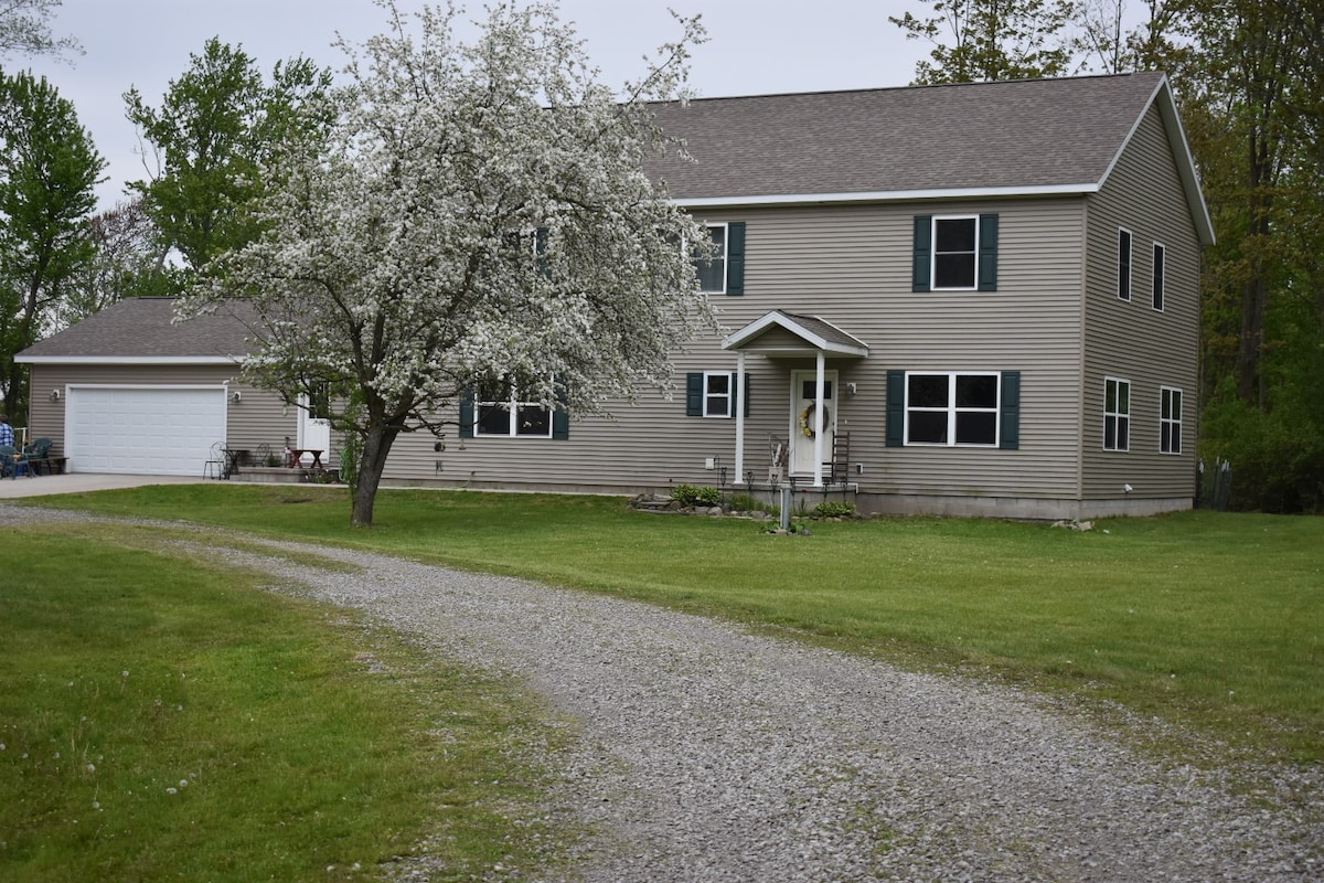 A spacious two-story house is framed by a gravel driveway, leading up to a welcoming entrance. An ornamental tree in bloom adds a touch of nature, while large windows reflect the surrounding greenery. A garage is positioned to the left of the structure.
