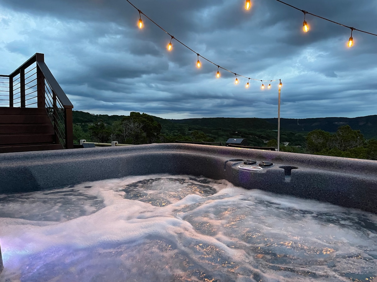 A hot tub filled with bubbling water is enclosed on a wooden deck, set against a backdrop of rolling hills under a cloudy sky. String lights overhead provide soft illumination, enhancing the serene ambiance of the outdoor space.