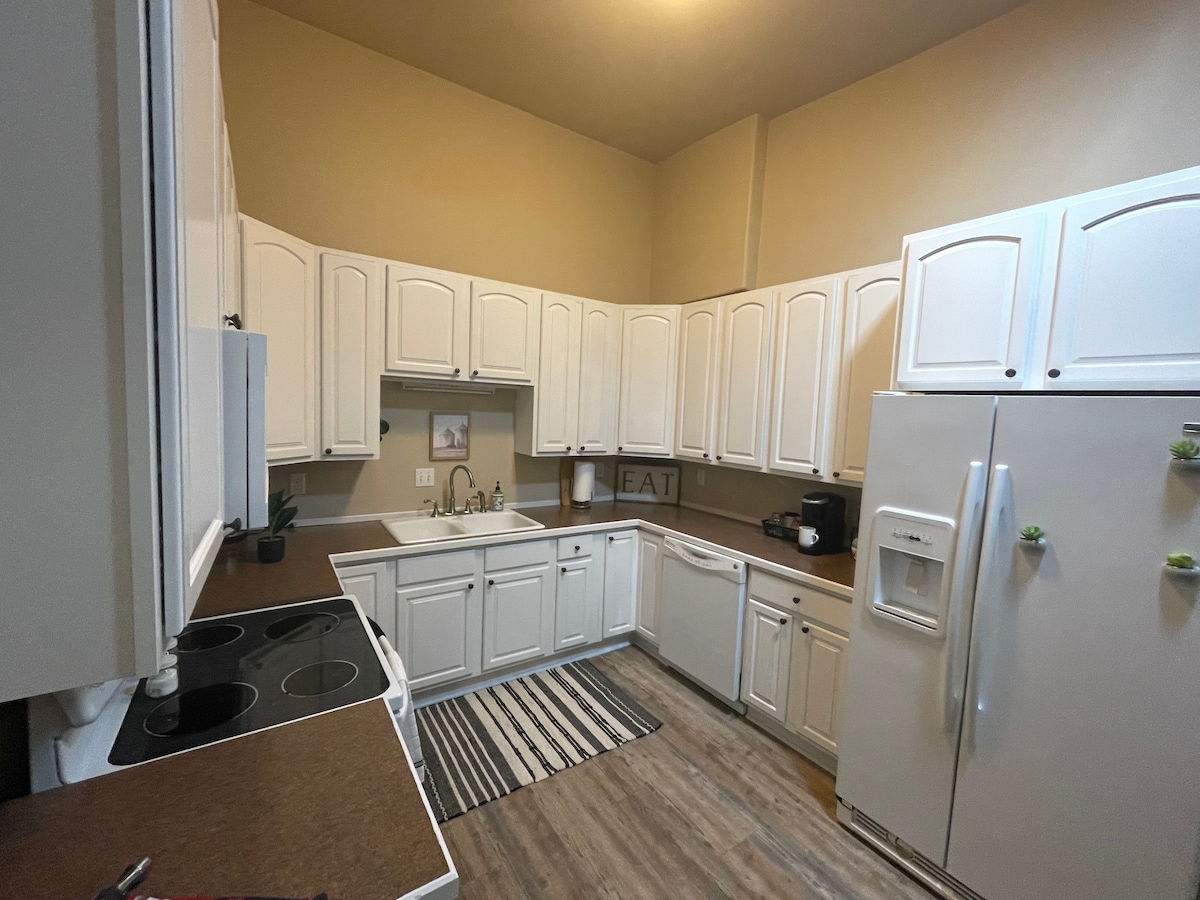 The kitchen features white cabinetry and a spacious countertop area. Equipped with a stove, oven, refrigerator, and coffee maker, it is designed for convenience. A striped rug adds texture to the floor, and a small wall art piece is visible above the sink.
