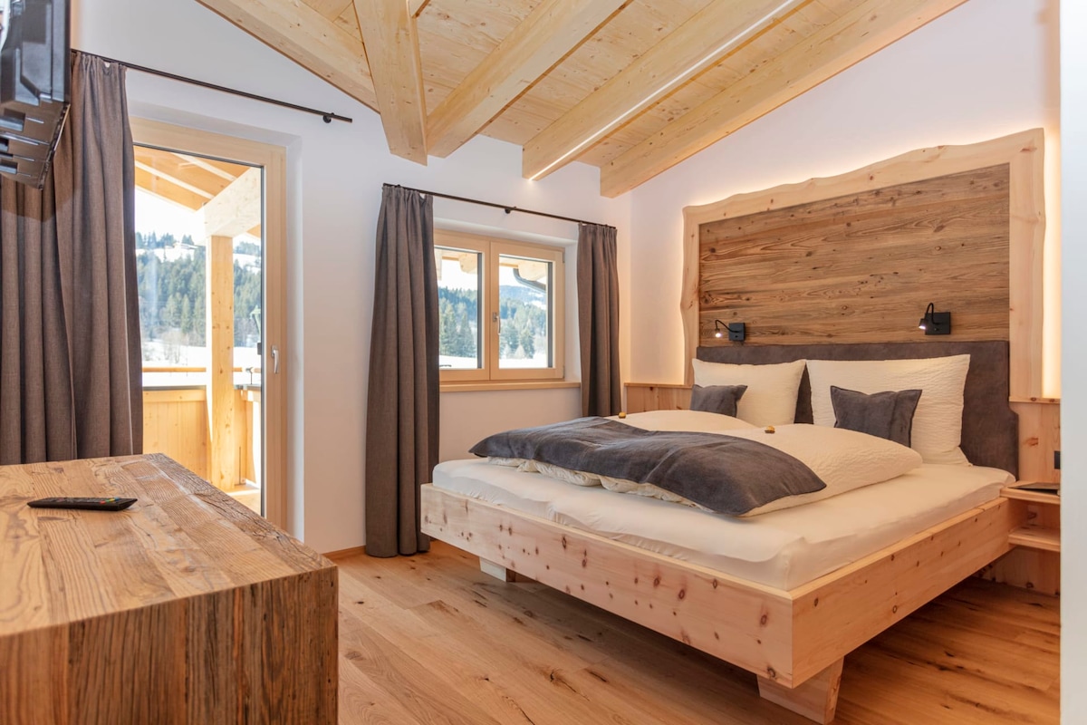 A cozy bedroom features a spacious bed with soft linens and a wooden headboard. Natural wood elements are present in the beams and furniture. Large windows with dark curtains offer views of the outdoors, while a balcony door provides access to fresh air.