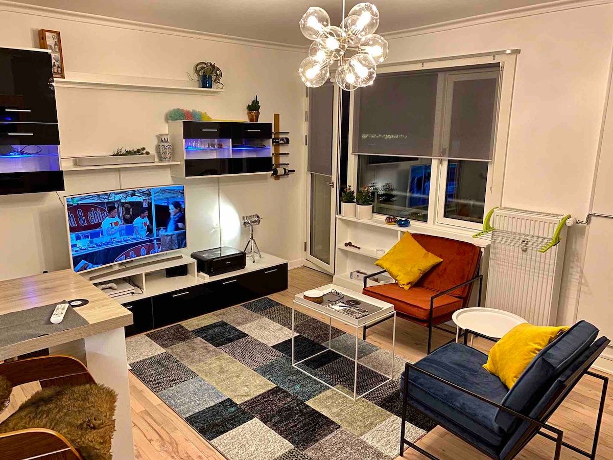 A modern living area features a patterned area rug, two comfortable accent chairs, and a sleek TV stand with a flat-screen television. A stylish light fixture hangs above, while large windows allow natural light to enter the space, creating a bright and inviting atmosphere.