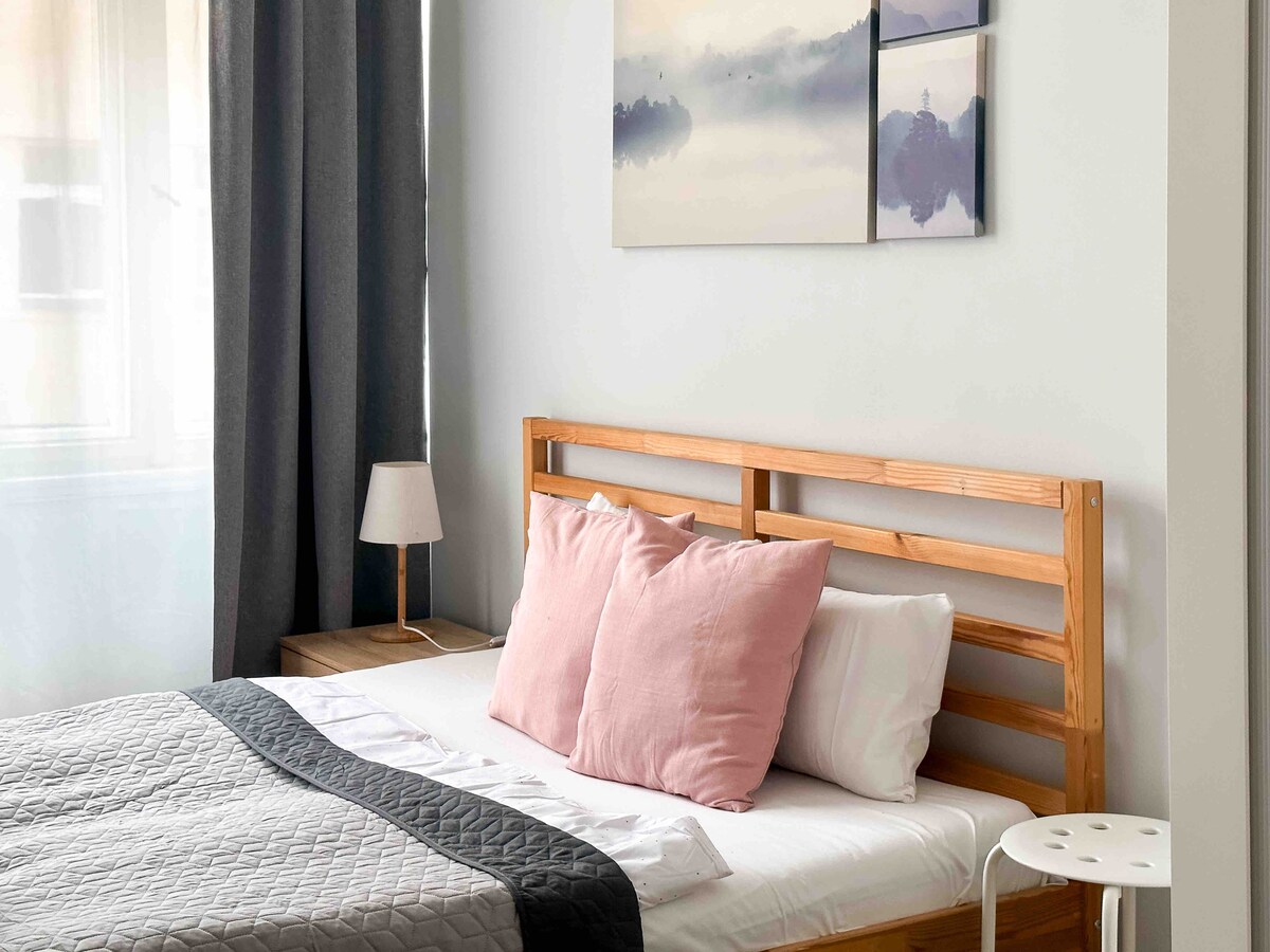 A bright bedroom features a spacious bed with soft pillows and an understated quilt. Two pastel art pieces decorate the wall above the bed. A small bedside lamp and a minimalistic stool complement the natural wood elements, while light filters through sheer curtains.