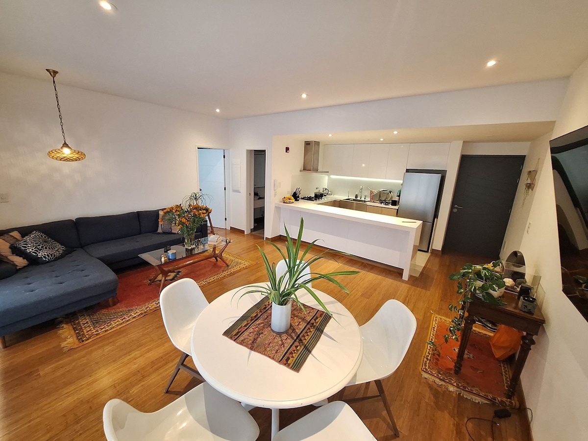 A modern open-concept living space is showcased, featuring a large sectional sofa and a circular dining table surrounded by white chairs. Natural light floods the room, highlighting decorative plants on the table and various ornamented rugs on the wooden floor.