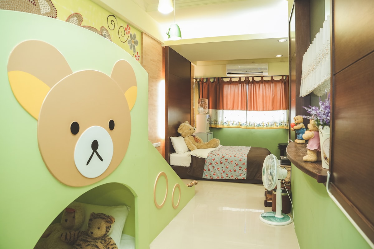 A children's themed room is featured, highlighting a playful bear-shaped slide and cozy sleeping areas. Plush toys are arranged on the bed and around the space, with warm orange curtains softly filtering light. A bedside table holds decorative items, contributing to a welcoming environment.