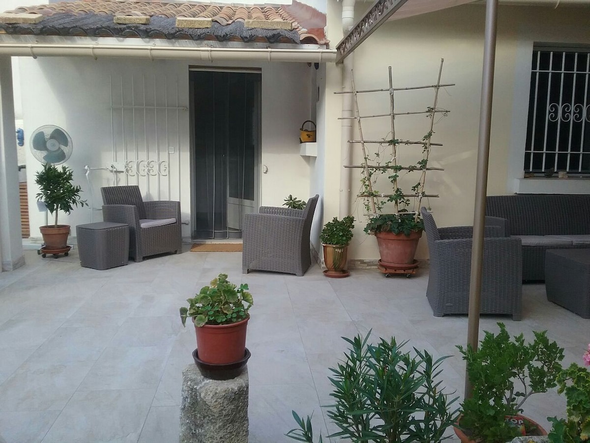 An outdoor space features several wicker seating arrangements with cushions, accompanied by potted plants. A shaded area is created by an overhead structure, while decorative greenery adds a fresh touch to the welcoming courtyard atmosphere.