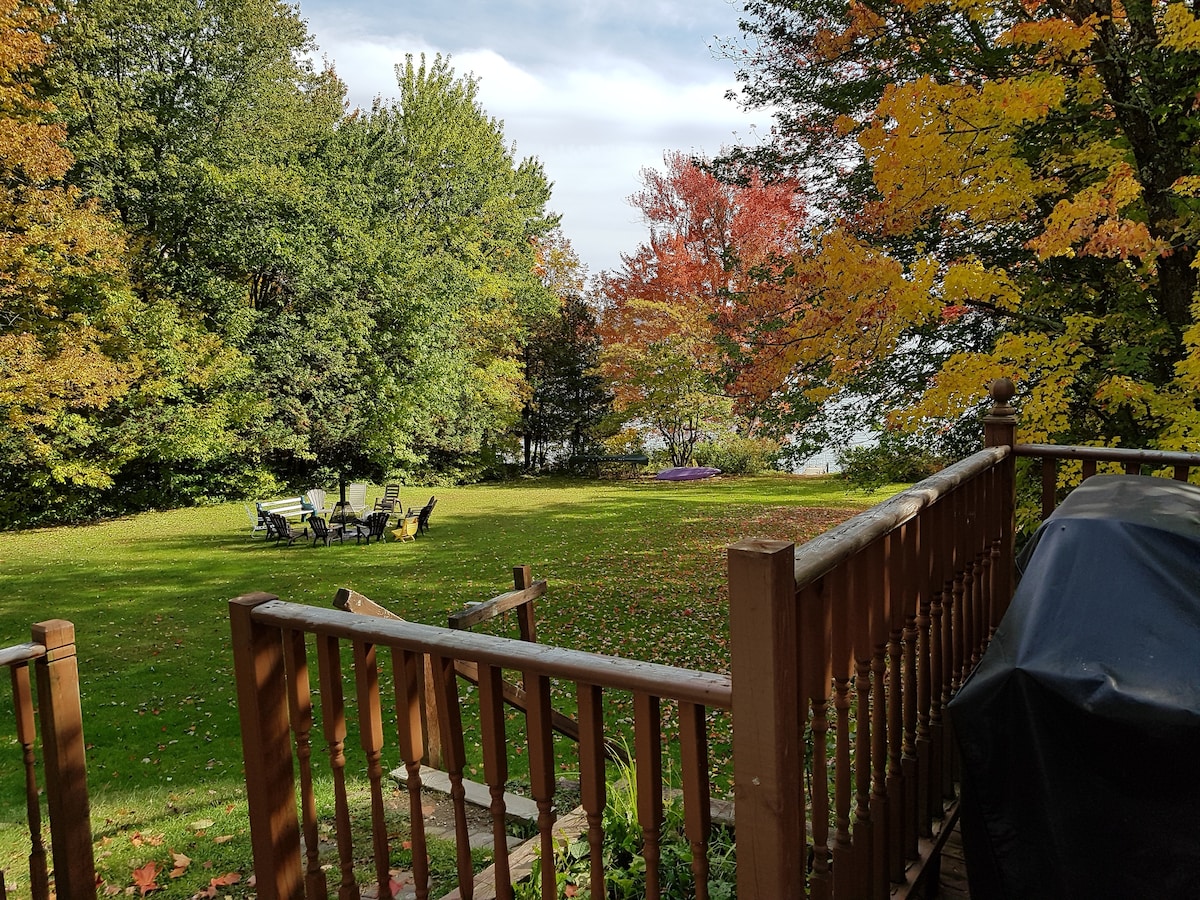 A spacious backyard features a well-maintained lawn bordered by a variety of trees displaying autumn colors. A seating area with chairs is arranged on the grass, offering a peaceful view of the nearby lake.