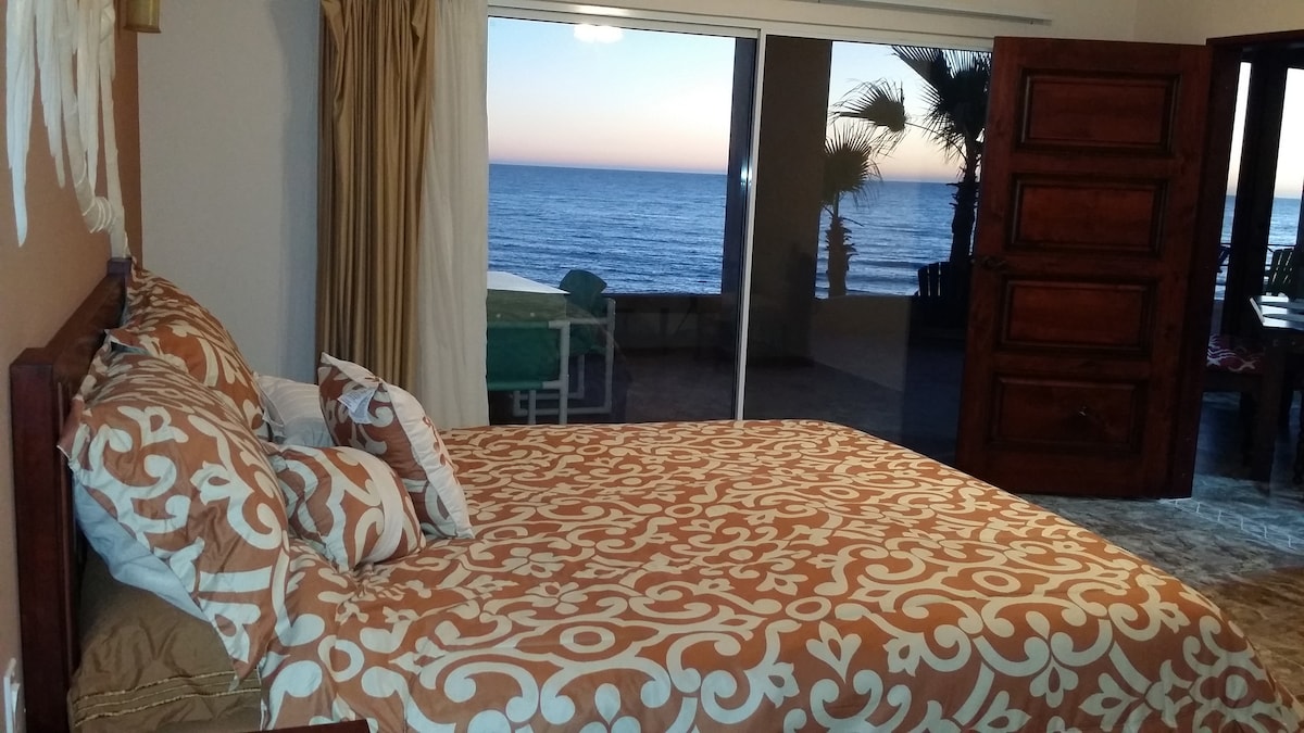 The bedroom features a spacious bed with an ornate orange and white patterned duvet. Large glass doors open to a view of the ocean and patio, with soft evening light illuminating the room. A palm tree silhouette is visible outside.
