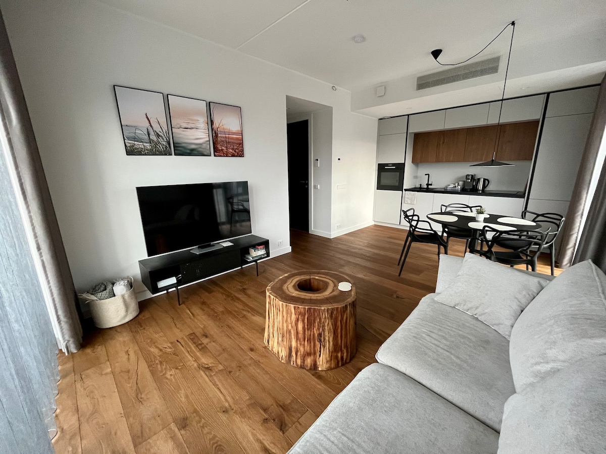 A modern living area is highlighted by a sleek television on a minimalistic stand. A round wooden coffee table is placed in front of a comfortable grey sofa. The dining area features a black dining table surrounded by matching chairs, with contemporary art on the wall above.