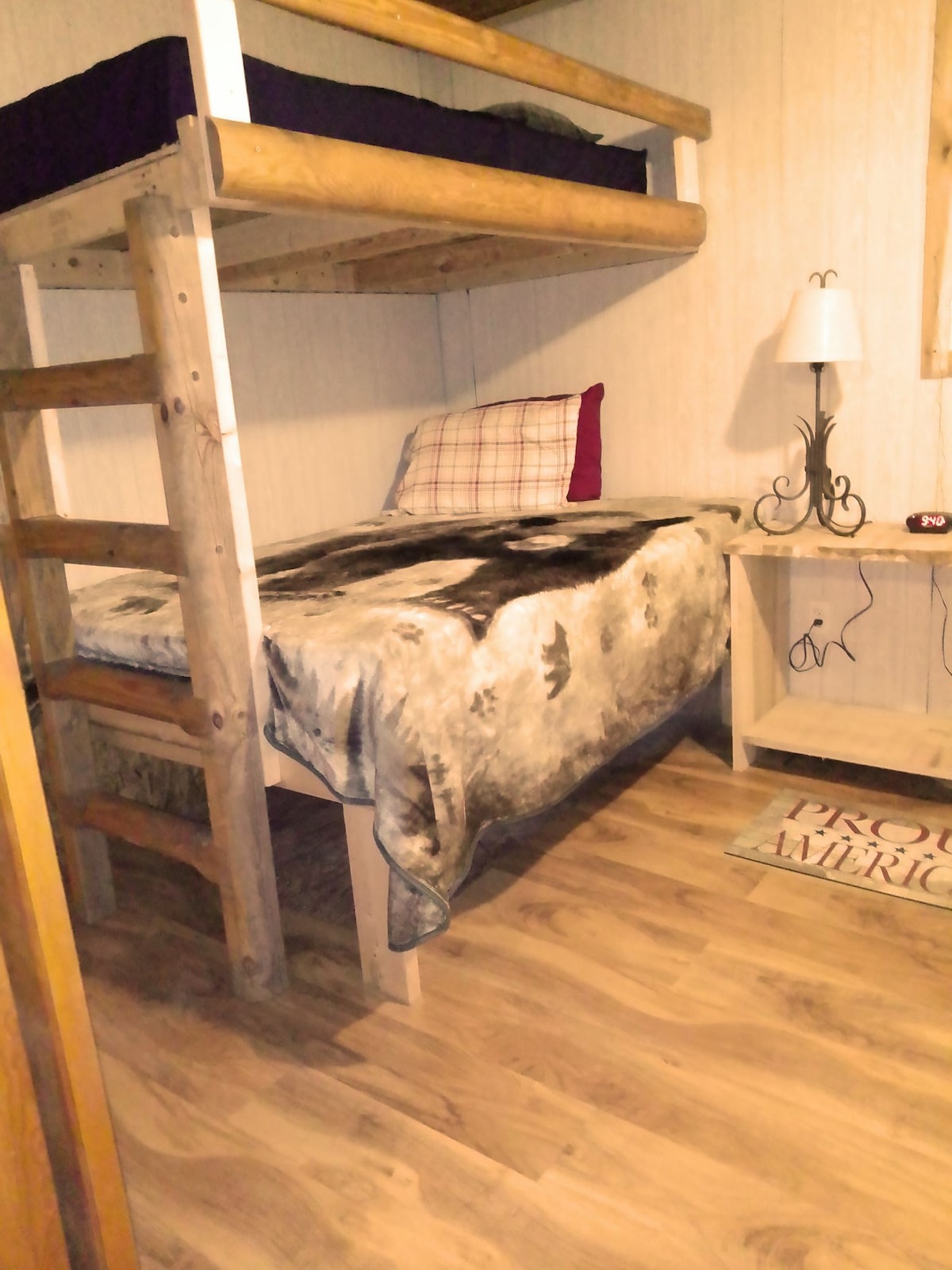 A wooden bunk bed is positioned against the wall, with a cozy lower sleeping area adorned with a patterned blanket. A small nightstand with a lamp is nearby, complementing the warm wooden floors that add to the rustic cabin ambiance.