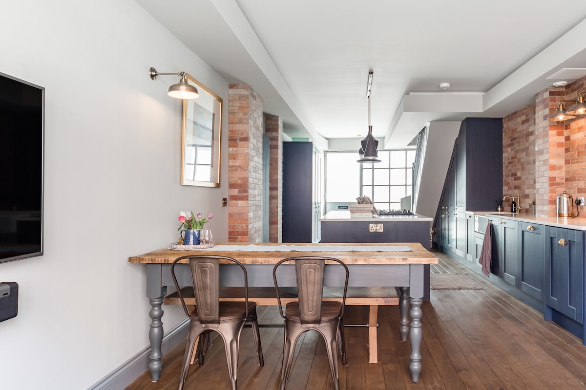 A bright kitchen and dining area is presented with a large wooden dining table flanked by metal chairs. Exposed brick walls are contrasted by modern cabinetry and an island featuring a central gas stove. Large windows provide ample natural light and a contemporary atmosphere.