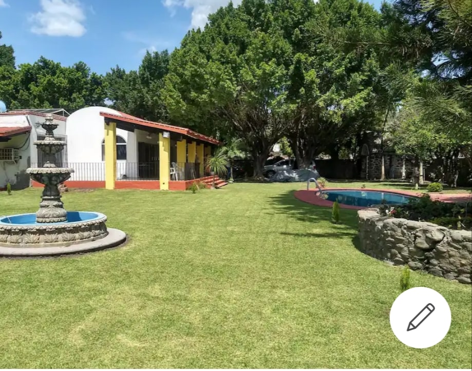 A spacious green garden is framed by tall trees, featuring a stone fountain at its center. Nearby, a pool is visible, with vibrant landscaping enhancing the serene atmosphere. Colorful buildings can be seen in the background, complementing the outdoor space.
