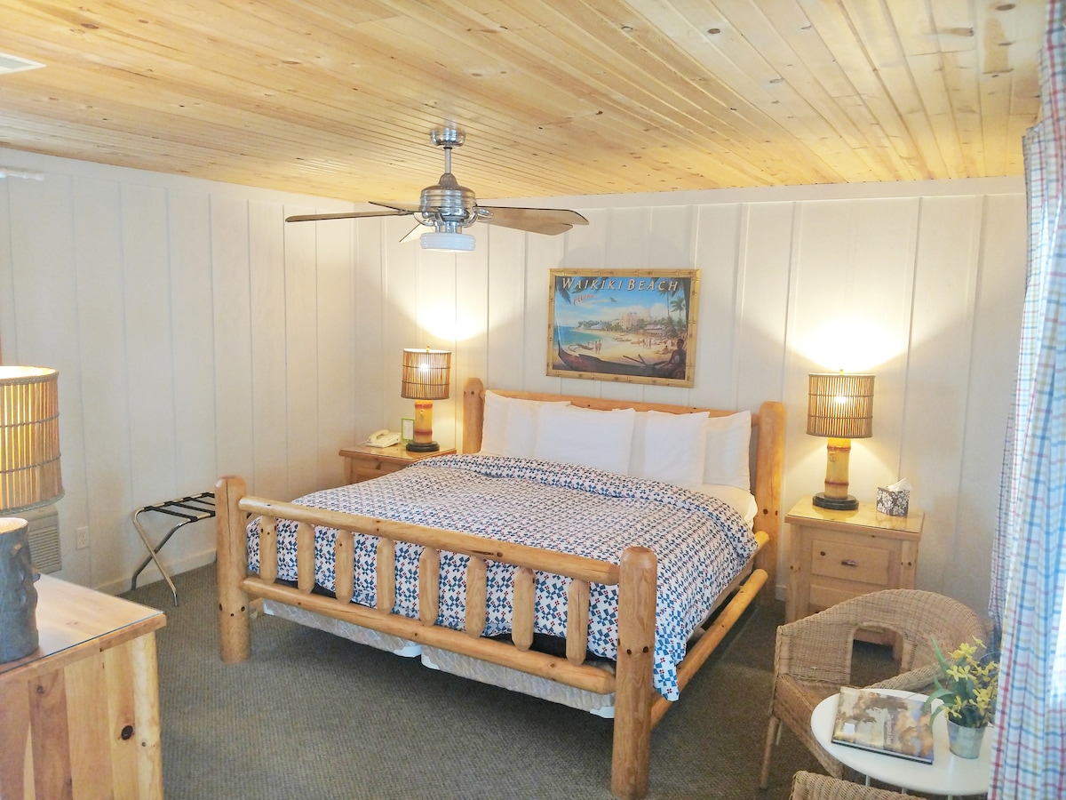 The sleeping area features a king bed with a patterned blanket atop a log frame. Flanking the bed are two wooden nightstands, each with a lamp. A ceiling fan is installed above, and a piece of artwork adorns the wall, creating a warm and cozy environment.