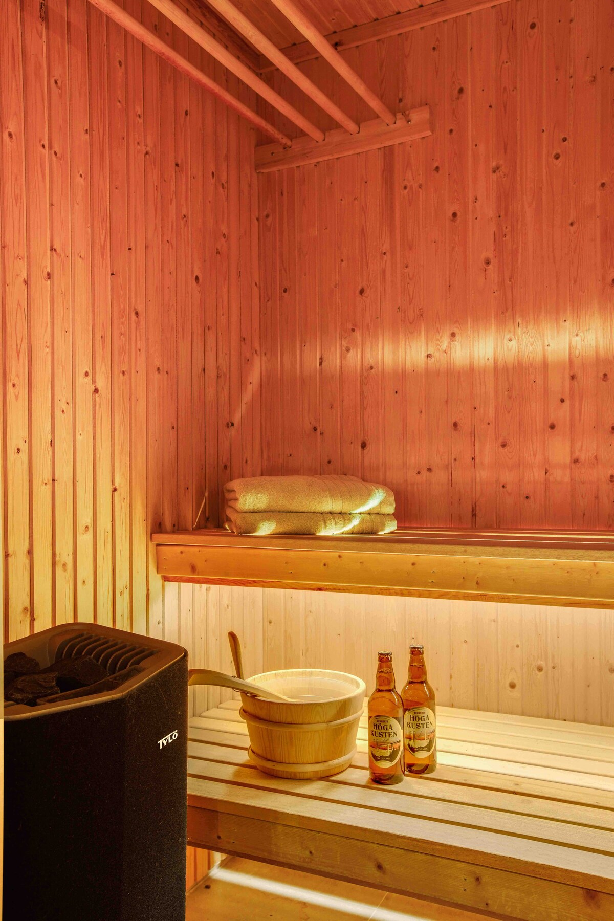 A wooden sauna features warm, ambient lighting, highlighting the smooth wooden panels of the walls and benches. A metal heater sits beside a traditional wooden bucket, accompanied by two bottles placed on the lower shelf. Soft towels are neatly arranged on the upper bench.