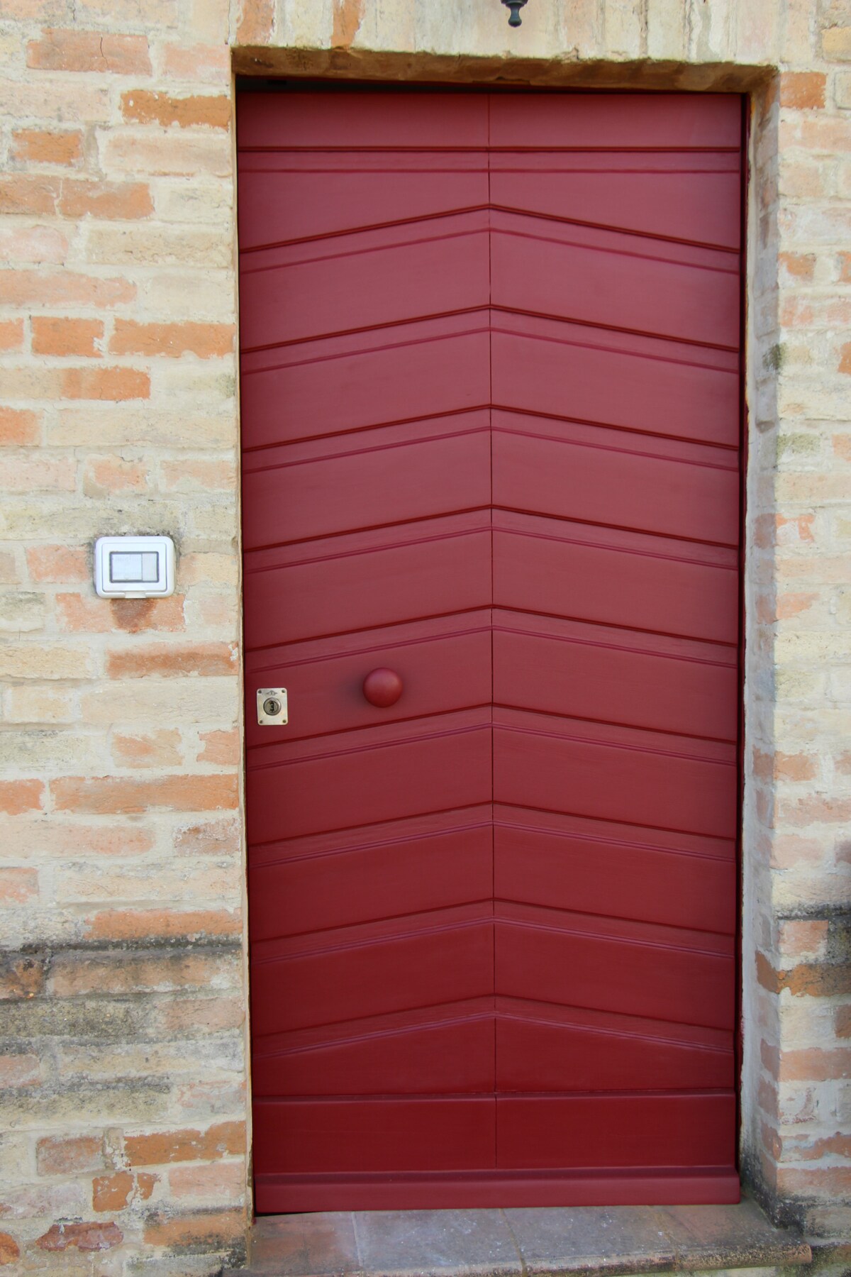 A striking red door with a chevron pattern stands against a textured brick wall. The door features a round handle and a small bell, inviting guests to enter. The earthy tones of the bricks complement the bold hue of the door, drawing attention to the entrance.