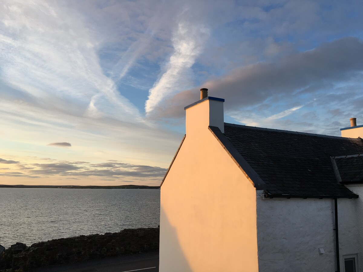 The image captures the corner of a white cottage, with a shingled roof, overlooking a calm body of water. The sky is filled with soft clouds during sunset, casting a warm glow onto the cottage's exterior and creating a serene atmosphere.