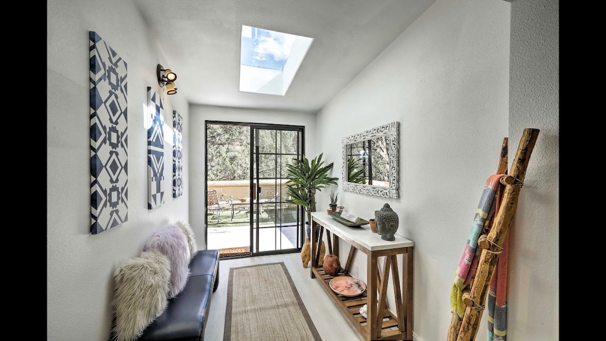 A bright entryway is highlighted by a large skylight and sliding glass doors leading to an outdoor space. A sleek black bench sits against one wall, complemented by decorative wall art. A console table displays decorative items, while a potted plant adds a touch of greenery.