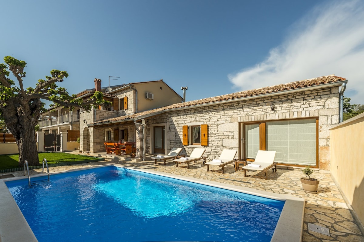 The exterior showcases a stone villa with a large outdoor swimming pool surrounded by sun loungers. The sunlit space includes a wooden dining set, and the villa’s classic architecture is complemented by a well-manicured garden, enhancing the tranquil atmosphere.