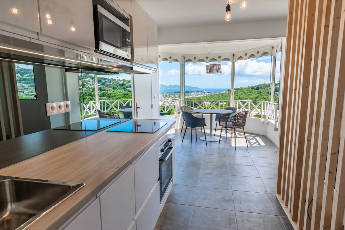 A modern kitchen features sleek cabinetry and a stainless steel oven, seamlessly connecting to a dining area with a round table and four chairs. Large windows offer a bright view of the surrounding greenery and bay, enhancing the airy ambiance.