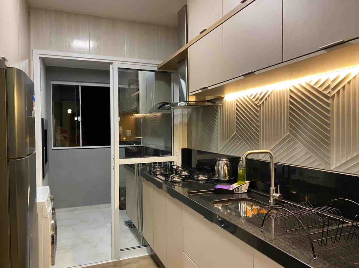 A modern kitchen features sleek cabinetry and a glossy black countertop. Cooking appliances, including a stove and kettle, are visible. A glass door provides access to an outdoor area, while soft lighting highlights the textured wall design.