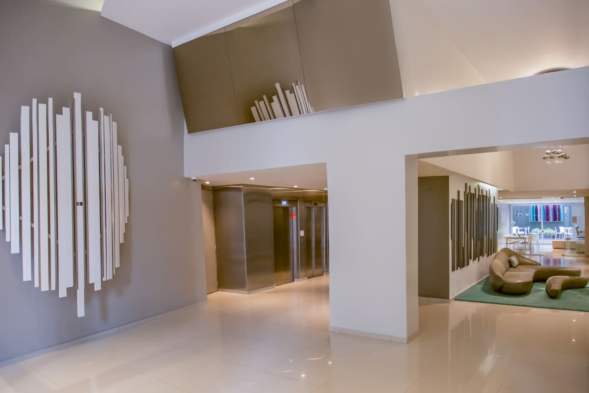 An expansive lobby area is highlighted by modern decor, featuring a large abstract wall art piece in light colors. Bright, reflective surfaces and soft lighting enhance the space, leading towards elevators and an inviting lounge area with curved seating, all set on glossy tile flooring.