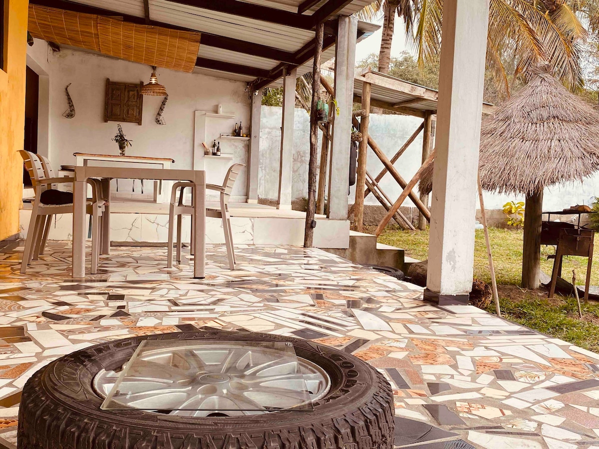 An outdoor terrace features a unique mosaic floor design. A table with chairs is situated under a covered area, providing a shaded space for relaxation. Nearby, a barbeque area is visible, along with a thatched roof structure, offering a tropical ambiance.