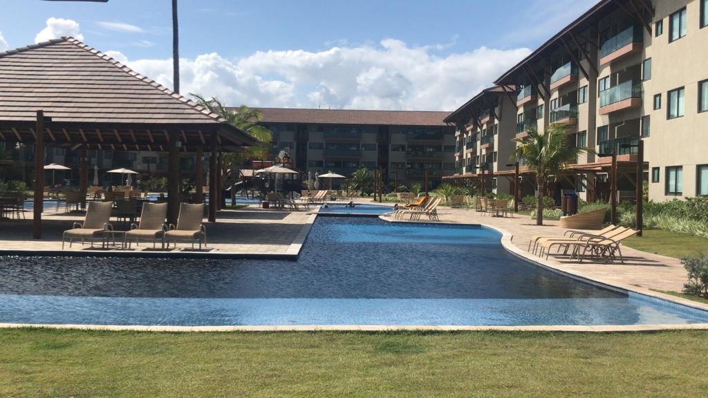An outdoor pool area is depicted, featuring a large swimming pool with a gradual slope. Lounge chairs are arranged along the water's edge, and umbrellas provide shade. Surrounding vegetation adds greenery, while a two-story building can be seen in the background against a clear sky.